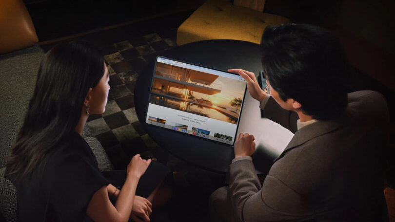 Two people sit at a round table, viewing architectural images on a large screen tablet or monitor in a dimly lit room.