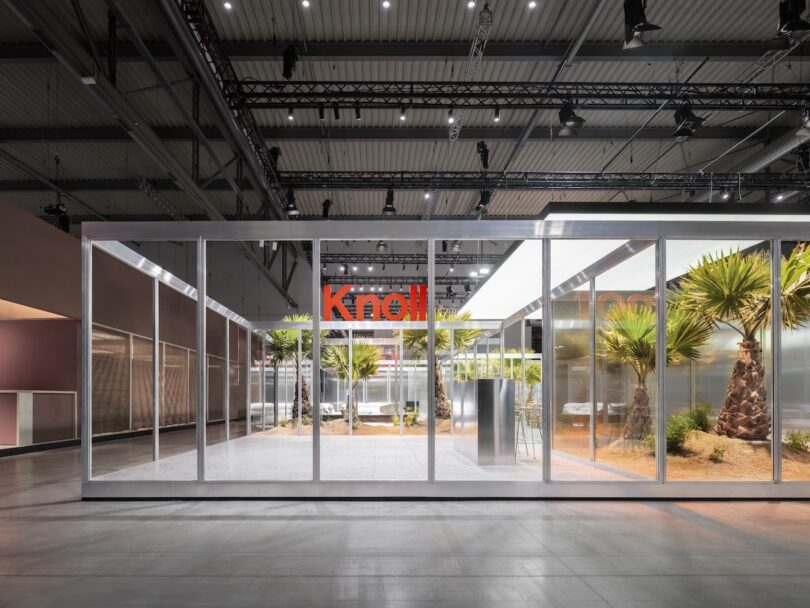 A modern glass-walled pavilion displays desert plants inside an industrial exhibition hall, with "Knoll" signage visible in the background