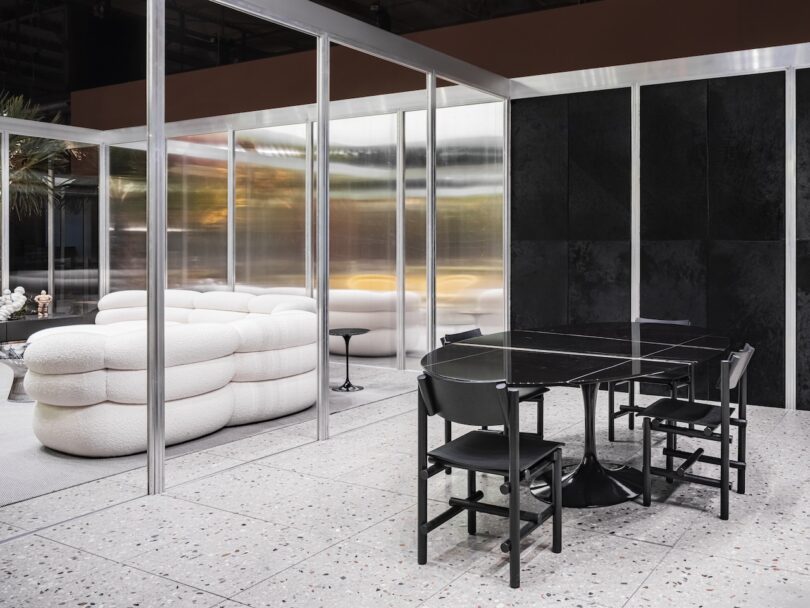 A modern interior features a black dining table with four chairs and a white curved sofa in a glass-partitioned space with terrazzo flooring and minimalist decor