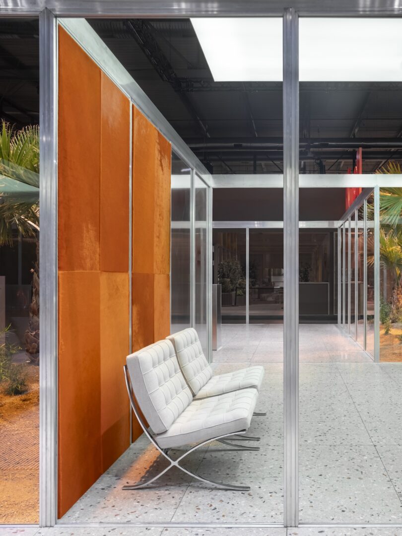 Modern interior with glass walls, terrazzo flooring, a white leather bench, and orange suede wall panels. Palm plants are visible outside the glass