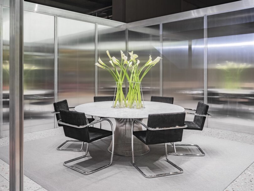 A modern round table with a metal base is surrounded by six black chairs. A tall vase with white calla lilies sits at the center. The space has glass walls and a neutral color scheme