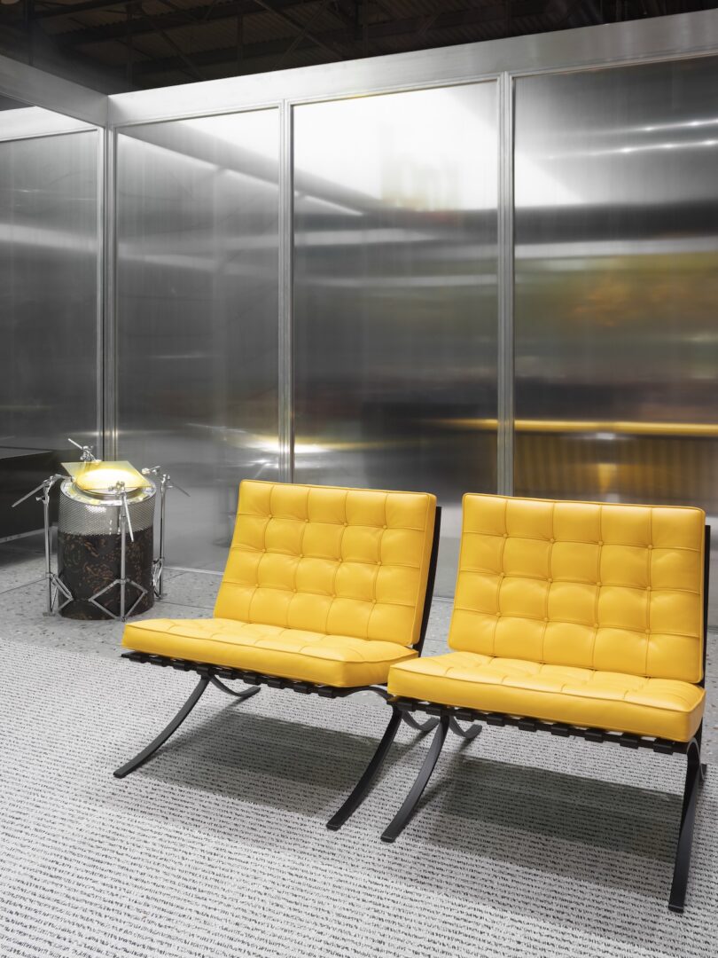 Two yellow tufted lounge chairs sit side by side on a gray carpet in front of frosted glass walls, with a drum set partially visible in the background