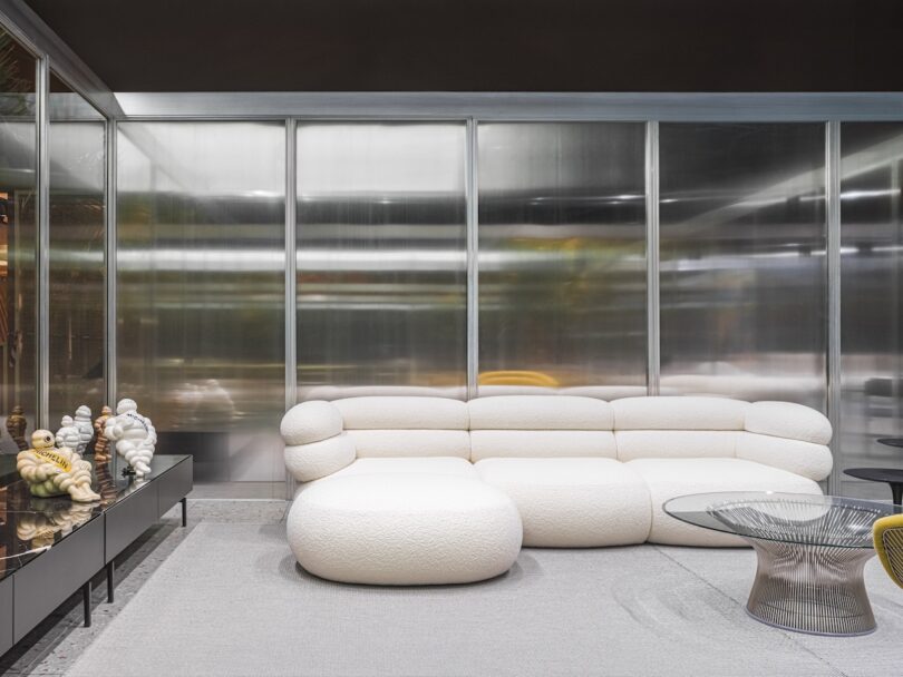 A modern living room with a white curved sectional sofa, glass coffee table, decorative sculptures on a sideboard, and frosted glass panel walls