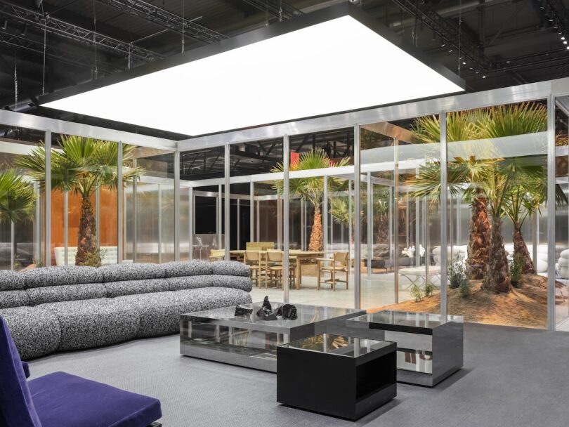 Modern living room with a gray sectional sofa, black coffee tables, and floor-to-ceiling glass walls revealing palm trees and patio seating outside. Large ceiling light overhead
