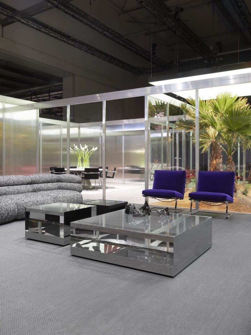 Modern lounge with glass coffee tables, two blue chairs, a patterned sofa, and an indoor garden visible through glass walls in the background