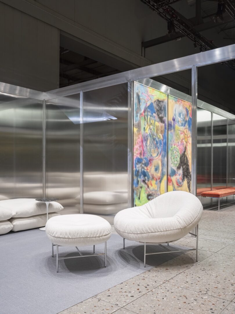 A modern lounge chair with matching ottoman on a gray rug, set in a minimalist room with metal walls, abstract art, and a bench in the background
