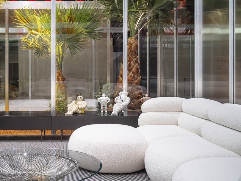 Modern lounge area with a curved white sofa, round glass coffee table, decorative sculptures, and large windows revealing palm trees outside