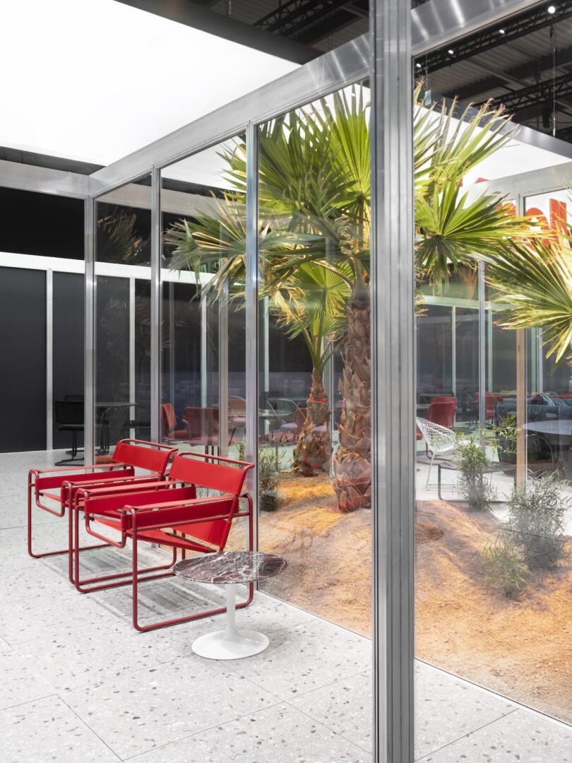 Two red modern chairs and a small round table sit inside a glass-walled room with palm trees and sandy landscaping outside