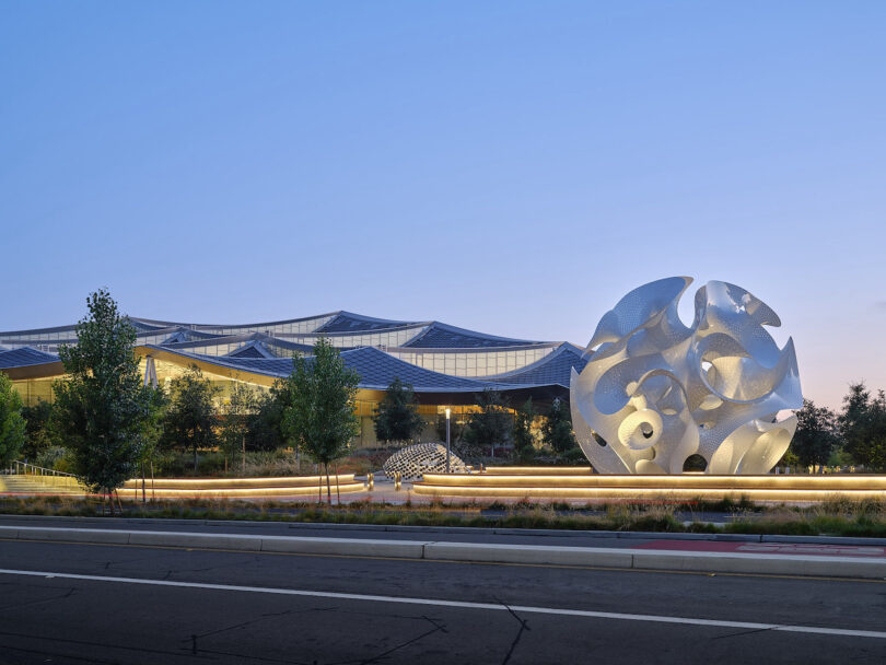 A modern building with a wavy roof stands behind a large white abstract sculpture and landscaping at dusk
