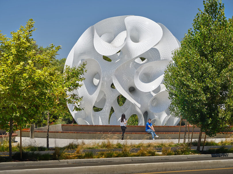 Two people sit near a large, white, abstract geometric sculpture surrounded by trees on a sunny day