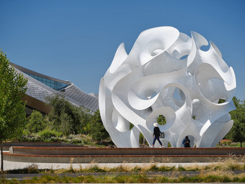 A large, abstract white sculpture with swirling shapes stands outdoors near modern buildings and landscaped greenery under a clear blue sky