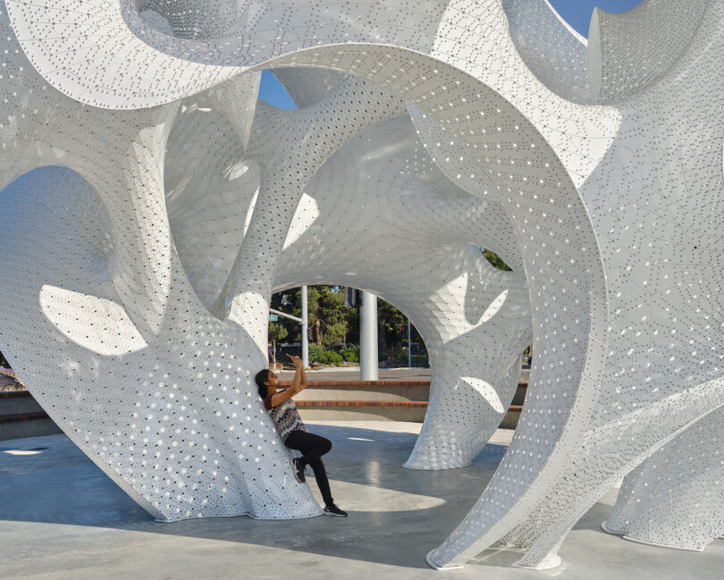 A person poses for a photo while sitting on a curved, white perforated architectural sculpture outdoors on a sunny day