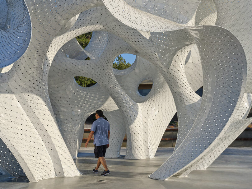 A man walks through a large, white, abstract metal sculpture with curved, perforated surfaces on a sunny day