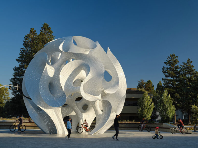 A large white abstract sculpture stands in an outdoor plaza as people walk and ride bicycles nearby under a clear blue sky