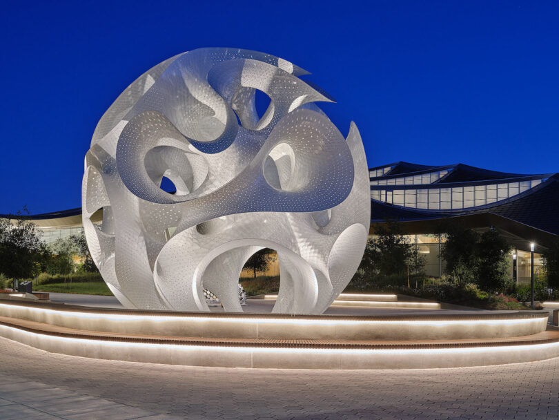 Large abstract metallic spherical sculpture with curved cutouts, illuminated at night, situated on a circular platform in an outdoor plaza