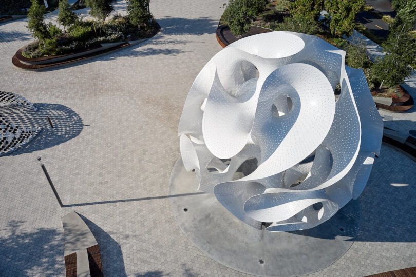 Aerial view of a large, abstract white metal sculpture with curved shapes in a landscaped plaza with trees and benches