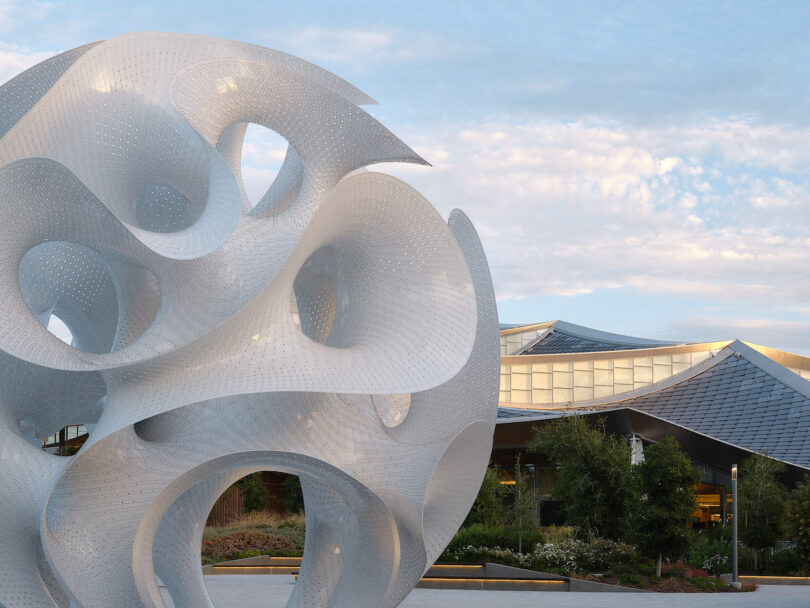 Large abstract white metal sculpture with swirling, perforated shapes stands outdoors in front of a modern building with glass and metal roof panels, under a partly cloudy sky