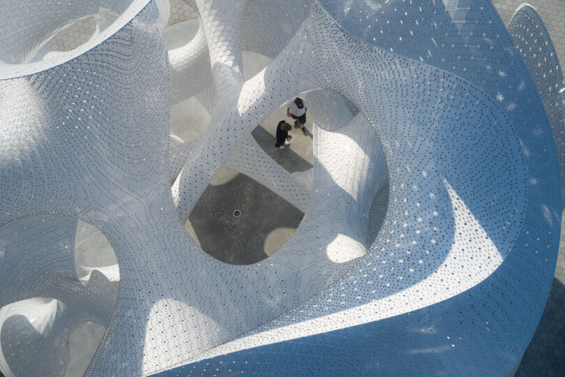A person and a child stand inside a large, white, perforated sculptural structure with curving, organic shapes, viewed from above
