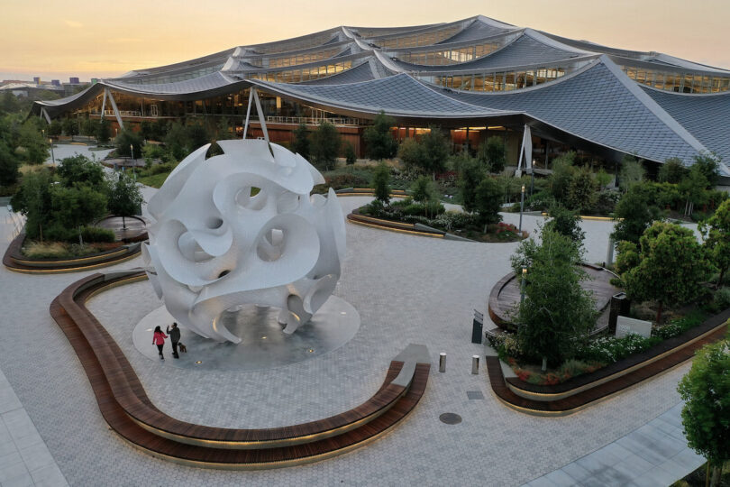 Modern architectural building with a wavy roof in the background, landscaped courtyard, and a large abstract white sculpture at the center; two people walk nearby.