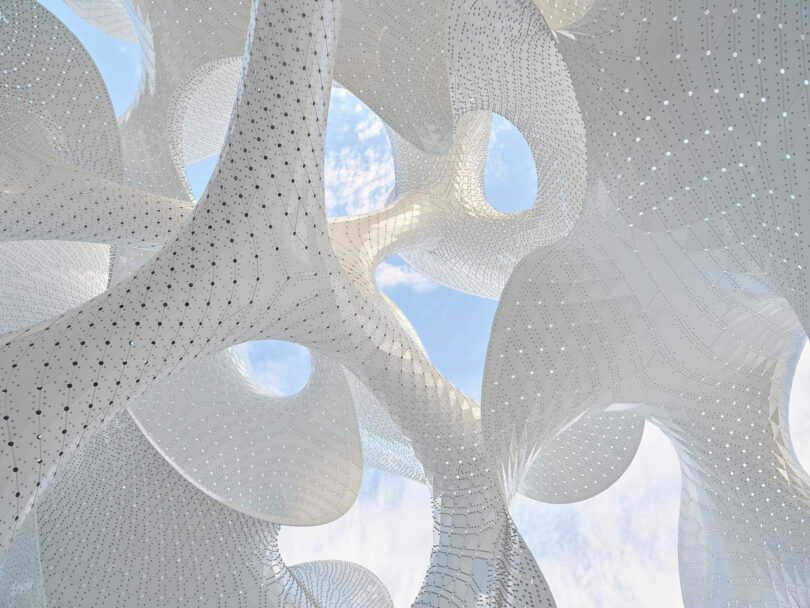 Abstract white sculptural structure with curved, perforated surfaces and large openings, set against a partly cloudy blue sky