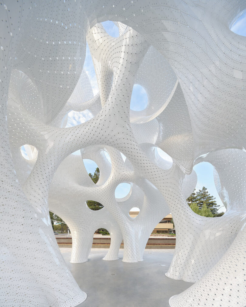 A large, white, sculptural pavilion with smooth, curving surfaces and circular openings, set outdoors with trees and sky visible through the structure