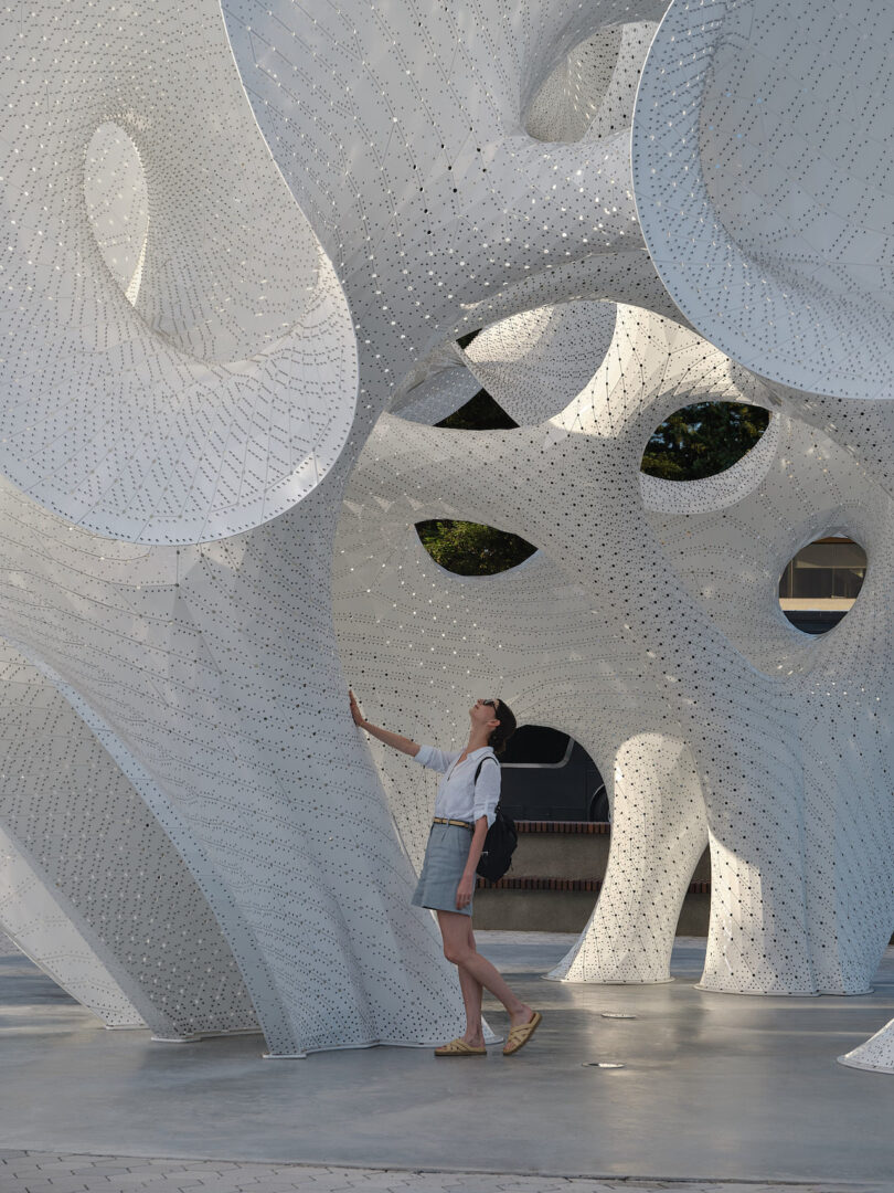 A person stands beneath a large, white, perforated abstract sculpture outdoors, looking up and reaching towards it