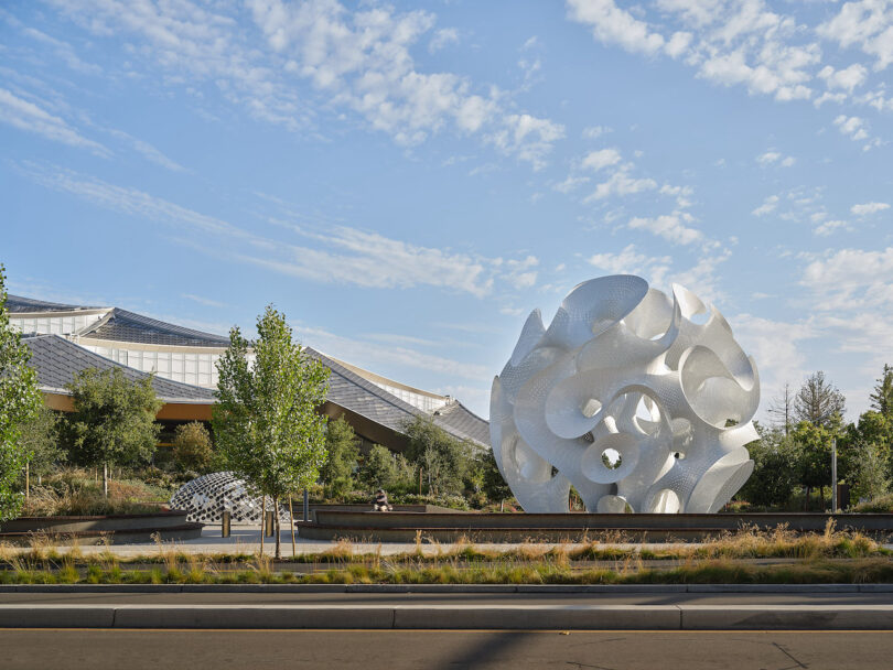 A large, intricate white metal sculpture stands in front of a modern building with a sloped glass roof, surrounded by trees and landscaped greenery under a partly cloudy sky