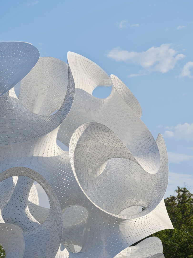 A close-up of a large, white, abstract outdoor sculpture with curved forms and perforated surfaces against a blue sky with scattered clouds