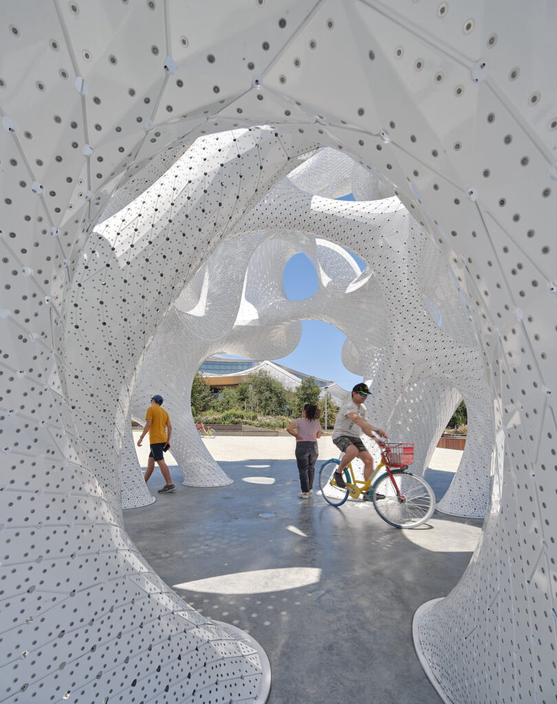 People walk and ride a bicycle through a large, white, perforated sculptural structure outdoors on a sunny day