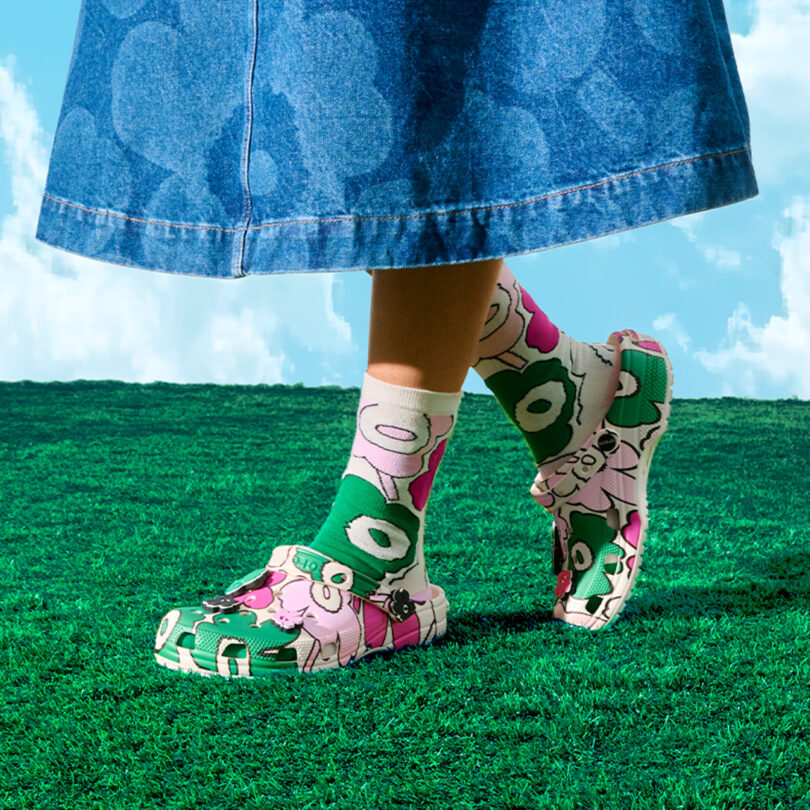 Person wearing a long denim skirt, colorful cartoon-patterned socks, and matching clogs stands on bright green grass with a blue sky background