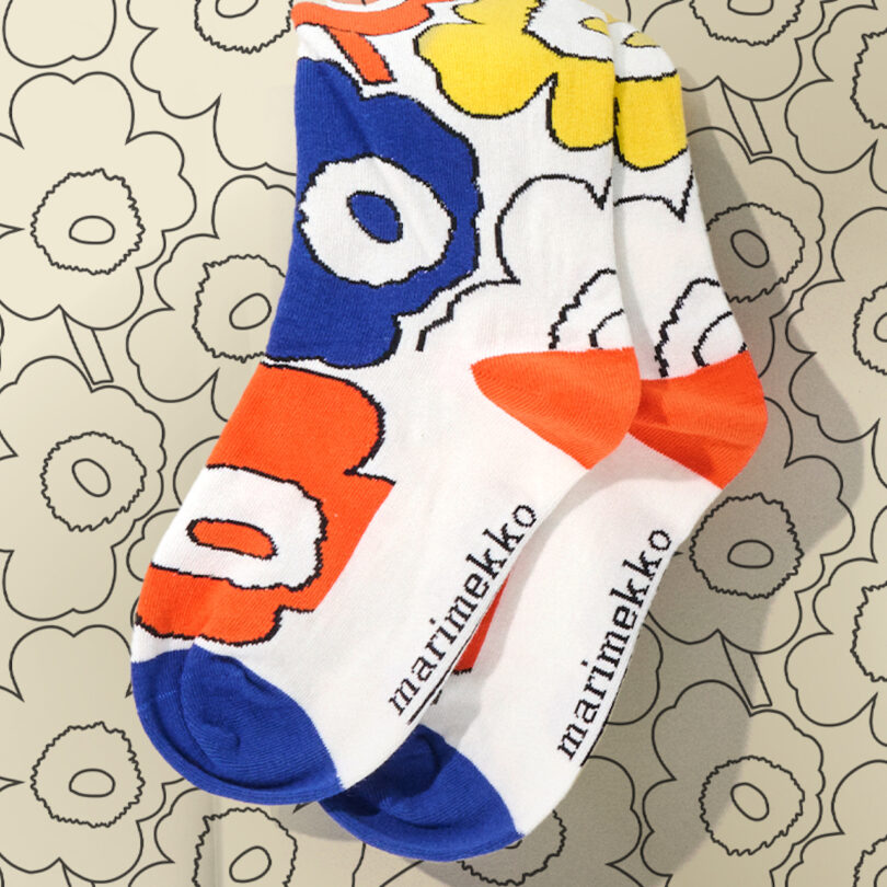 A pair of colorful Marimekko socks with a bold floral pattern in blue, red, yellow, and white, placed on a matching floral patterned background