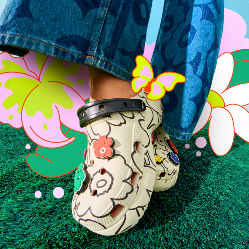 A person wears cream-colored Crocs with floral patterns and charms, standing on green grass with colorful floral and butterfly graphics in the background