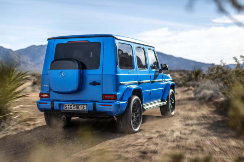 A bright blue Mercedes-Benz G580 G-Class SUV drives on a dusty desert trail with mountains and dry vegetation in the background.