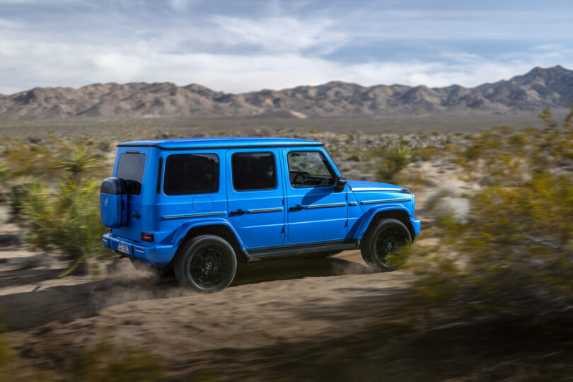 A blue Mercedes-Benz G-Class G580 SUV drives off-road through a desert landscape with mountains in the background.