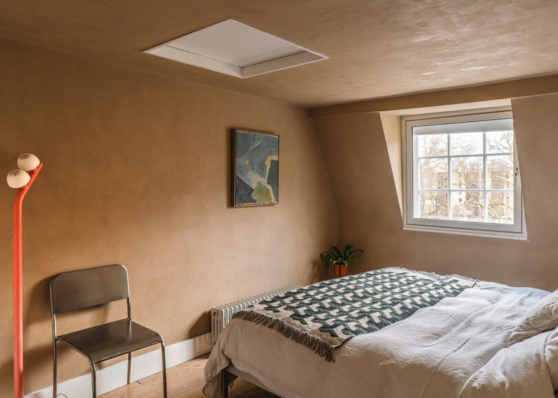 A simple bedroom with tan walls, a window, a bed with a patterned blanket, a metal chair, a plant on a radiator, a wall art piece, and an orange floor lamp with two bulbs