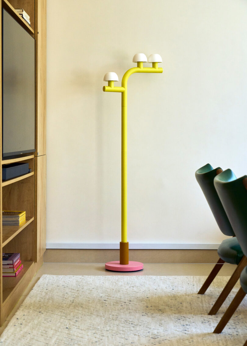 A modern yellow floor lamp with three white dome-shaped lights stands in the corner beside a wooden shelf and green chairs on a beige rug