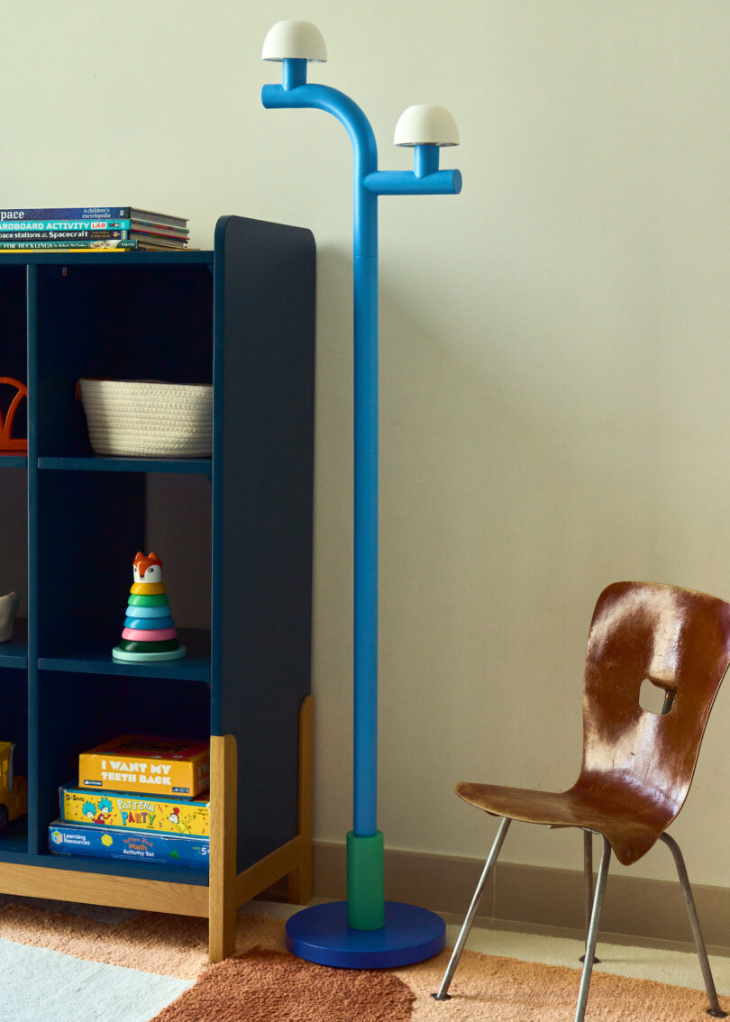 A blue floor lamp with two white dome lights stands beside a wooden chair and a shelf holding books and toys against a beige wall