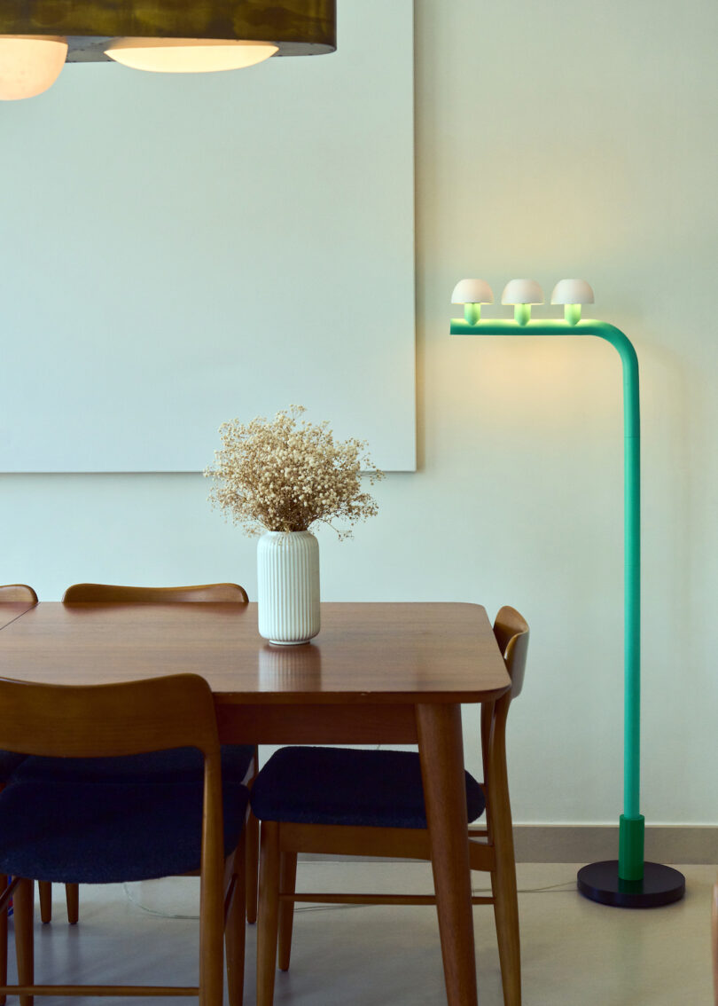 A wooden dining table with four chairs, a white vase with dried flowers, and a modern green floor lamp with three bulbs beside a blank wall