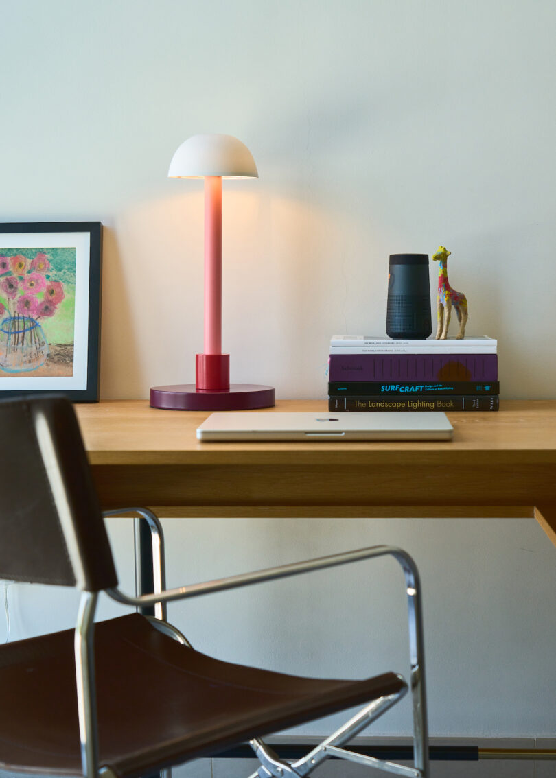 A wooden desk with a chair, a closed laptop, a stack of books, a lamp, a framed picture, a small figurine, and a cup against a plain wall