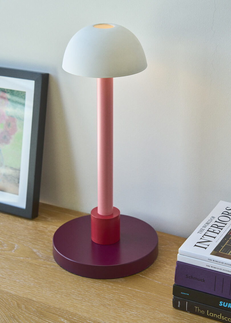 A modern table lamp with a white dome shade and pink base sits on a wooden surface next to books and a framed picture