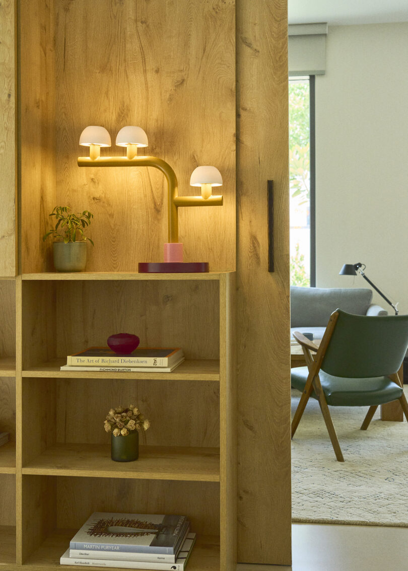 A wooden shelving unit with books, a small plant, dried flowers, and a modern brass lamp with three white globes, in a living space with a chair and window in the background