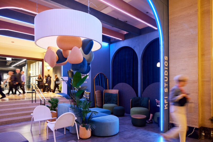 Modern lobby area with colorful, abstract light fixture, contemporary seating, blue accent walls, and people walking by; sign reads "KFI Studios"