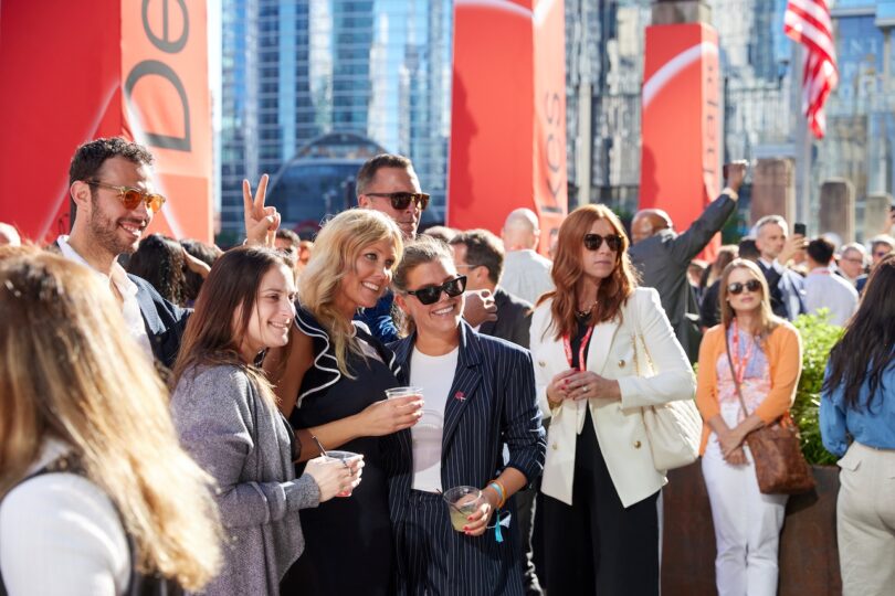 A group of people pose for a photo and socialize outdoors at a city event, with tall buildings and red banners in the background