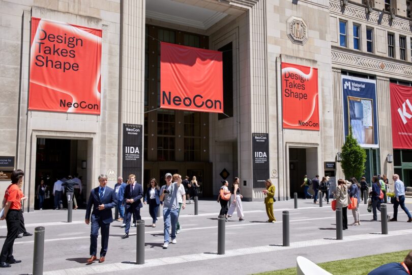 People walk outside a large building with red banners reading "Design Takes Shape" and "NeoCon" during a public event