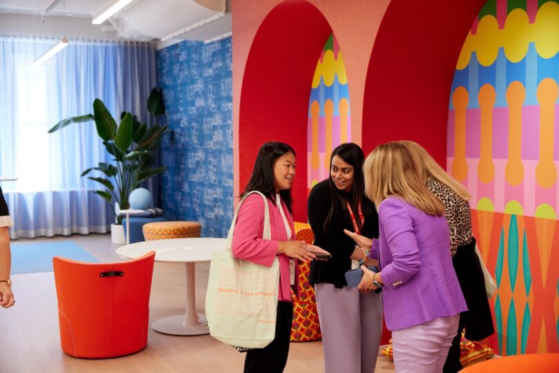 Four women stand and chat in a colorful, modern office space with bold wall patterns, bright furniture, and plants visible in the background