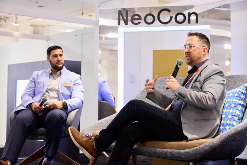 Two men in suits participate in a discussion on stage at NeoCon; one speaks into a microphone while the other listens attentively