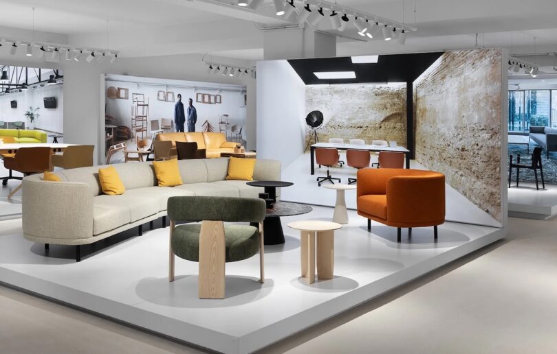 A modern showroom displays contemporary sofas, armchairs, and tables in neutral and orange tones, set on a raised platform with large interior photos in the background