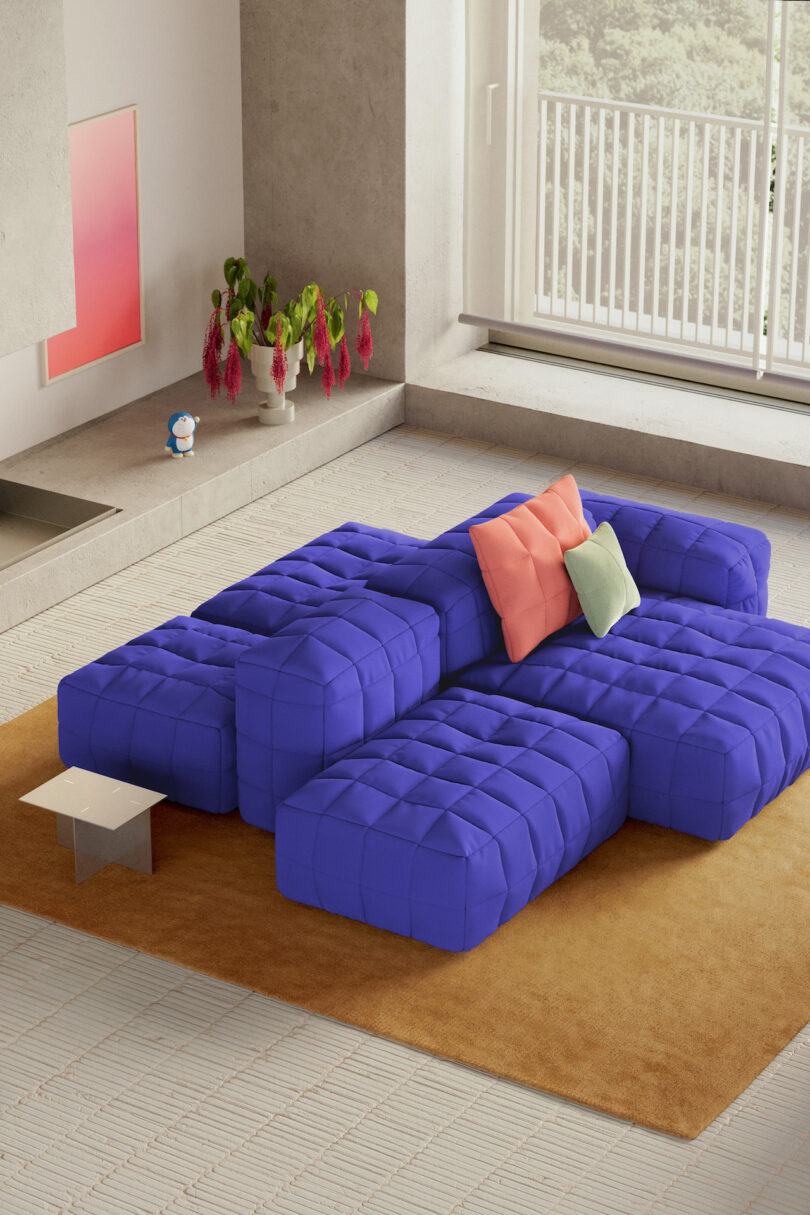 Modern living room with large windows, a blue modular sofa, pink and green cushions, brown rug, potted plant, and cityscape view in the background