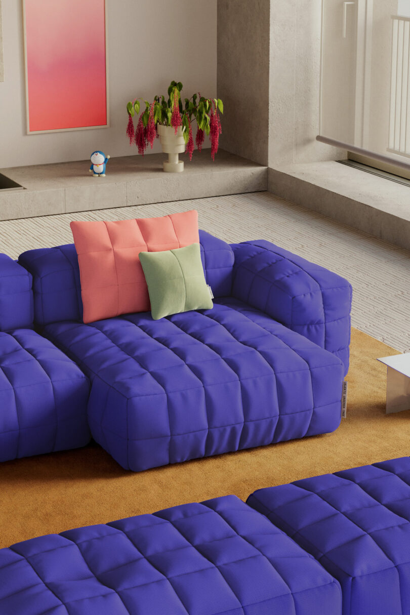 A modern living room with a blue tufted sofa, coral and green pillows, a yellow rug, a potted plant, abstract wall art, and a small decorative figurine on a ledge
