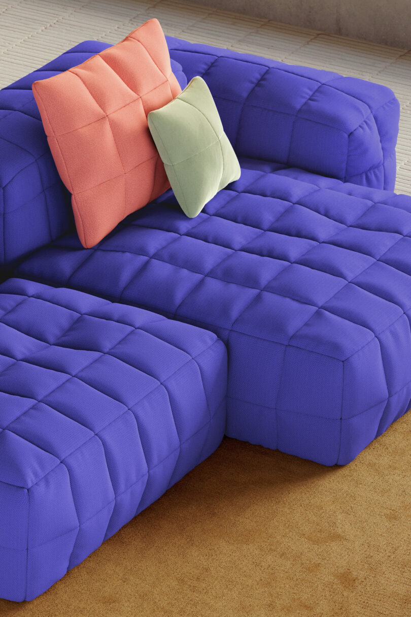 A modular sofa with quilted, bright blue cushions sits on a brown carpet, topped with one pink and one pale green pillow